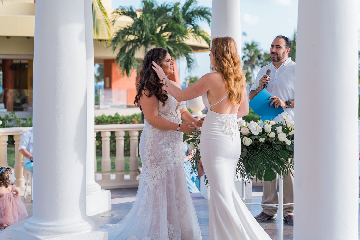 Two brides sharing an emotional moment at a beachside wedding gazebo ceremony with white columns.