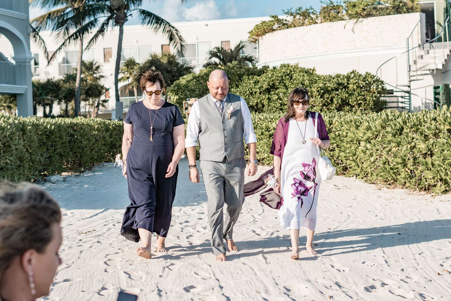 Groom in gray vest walks barefoot on sandy beach path escorted by two female guests.