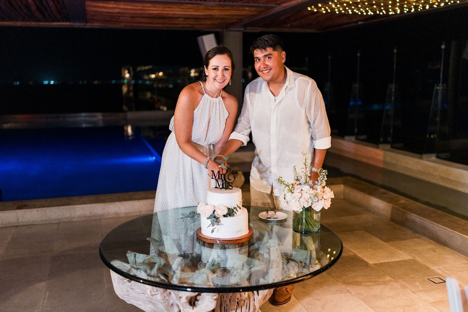 Bride and groom cut wedding cake together by poolside at night celebration.