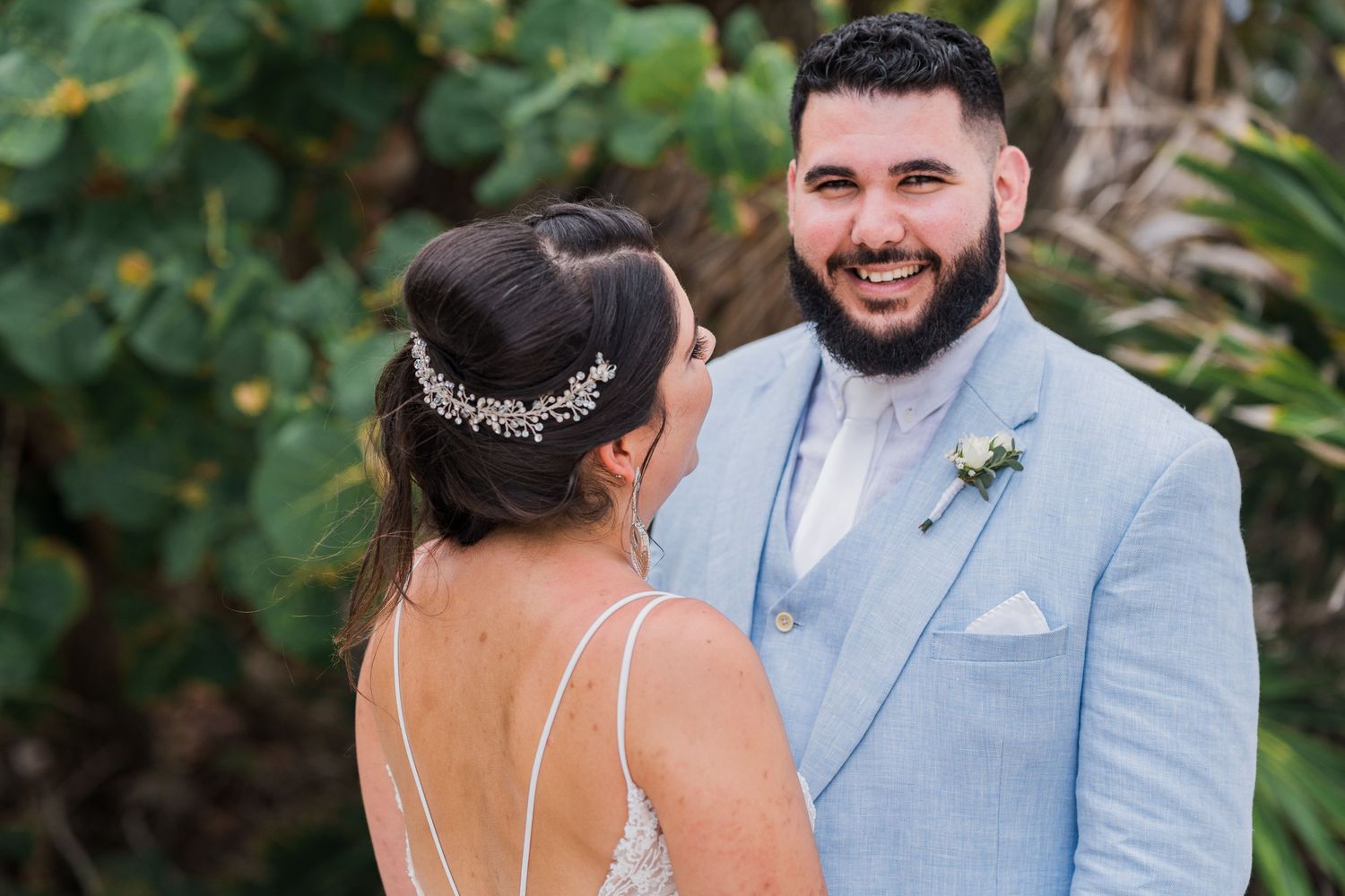 Smiling groom in a light blue suit looking at bride with tropical greenery in the background.