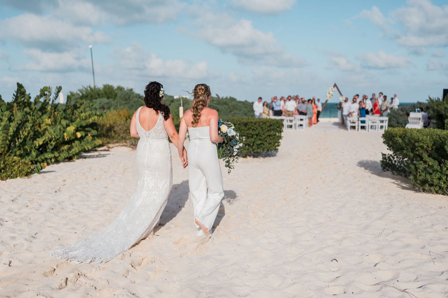 Two brides in white walk hand in hand along a sandy beach toward their wedding ceremony.
