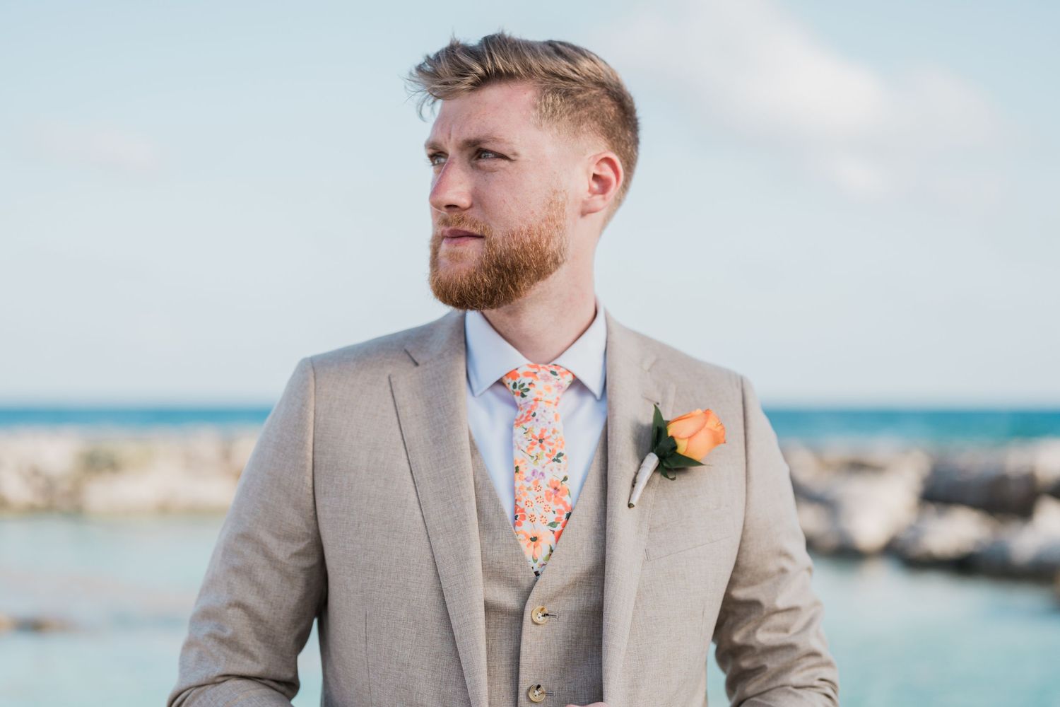 Handsome groom in a tan suit with floral tie and boutonniere gazes into the distance at an oceanside wedding.