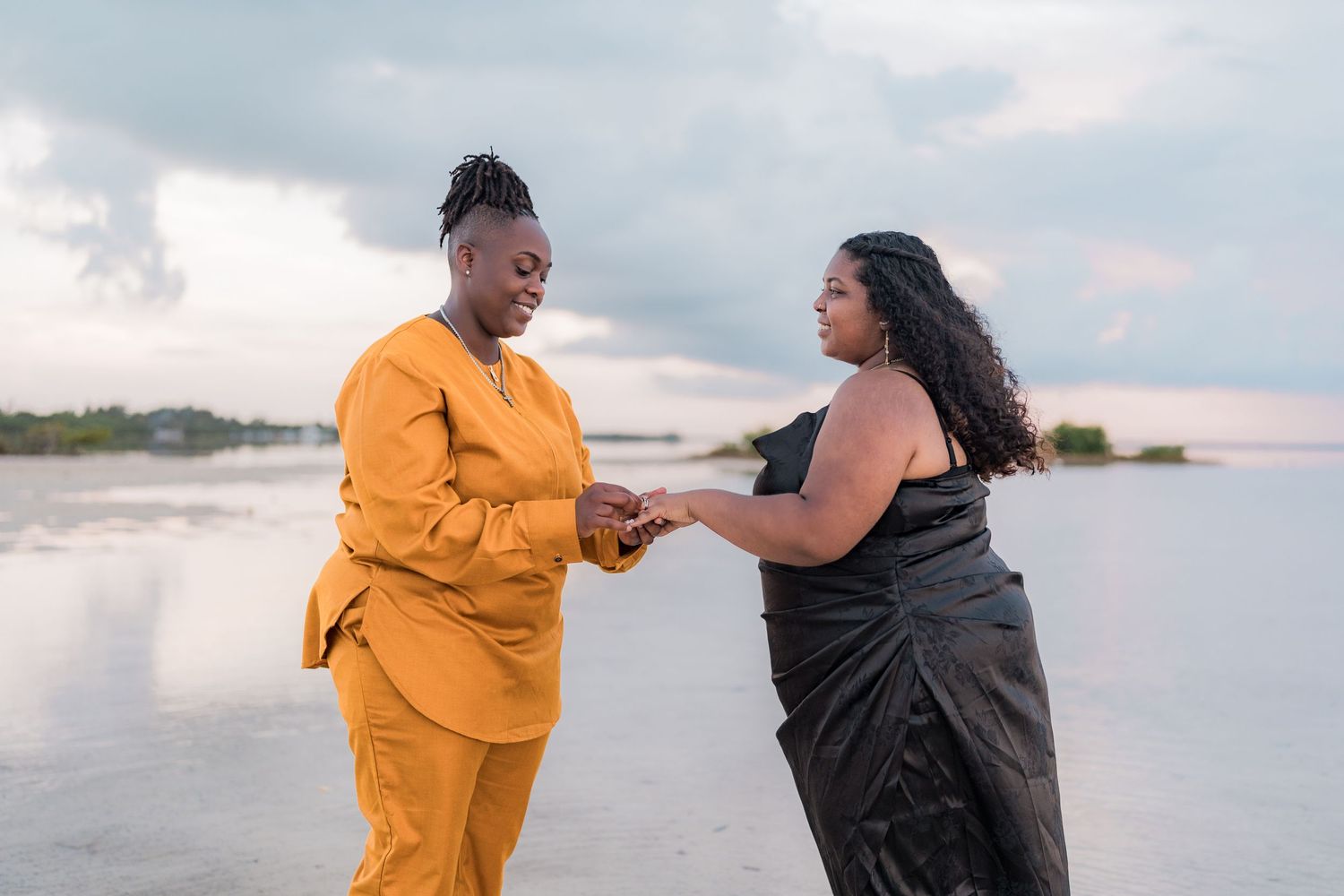 Two Black women holding hands on beach, one in orange suit and one in black dress, smiling.