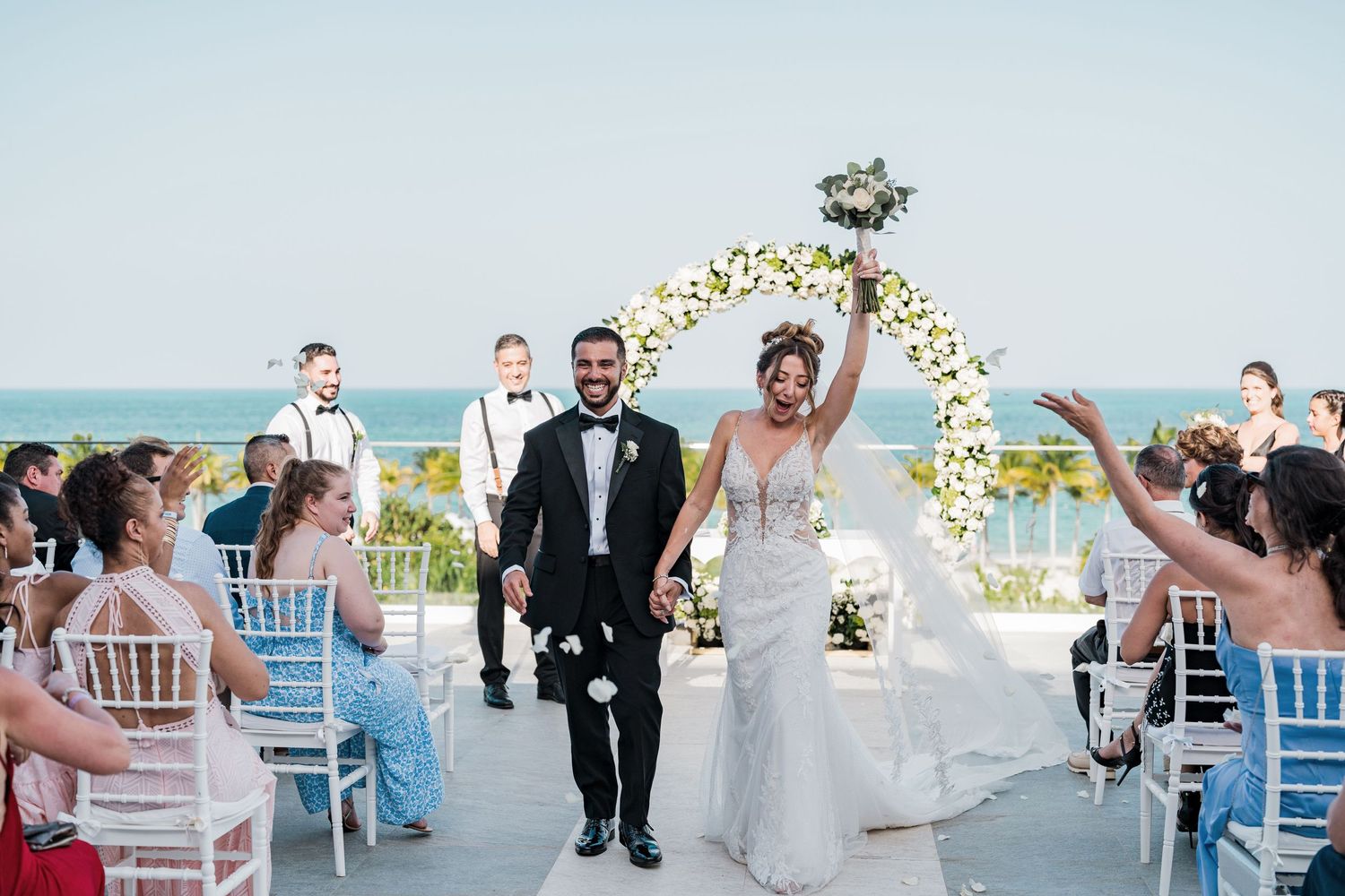 A radiant bride raises her bouquet in celebration as she and her groom exit their oceanfront ceremony.