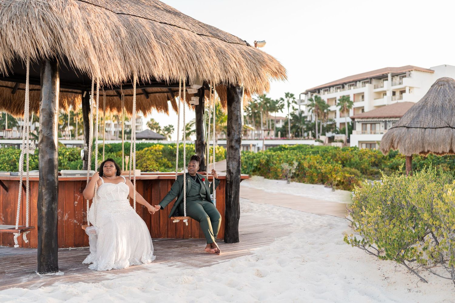 Bride and groom sit on wooden beach swings under a thatched palapa at a tropical resort wedding venue.