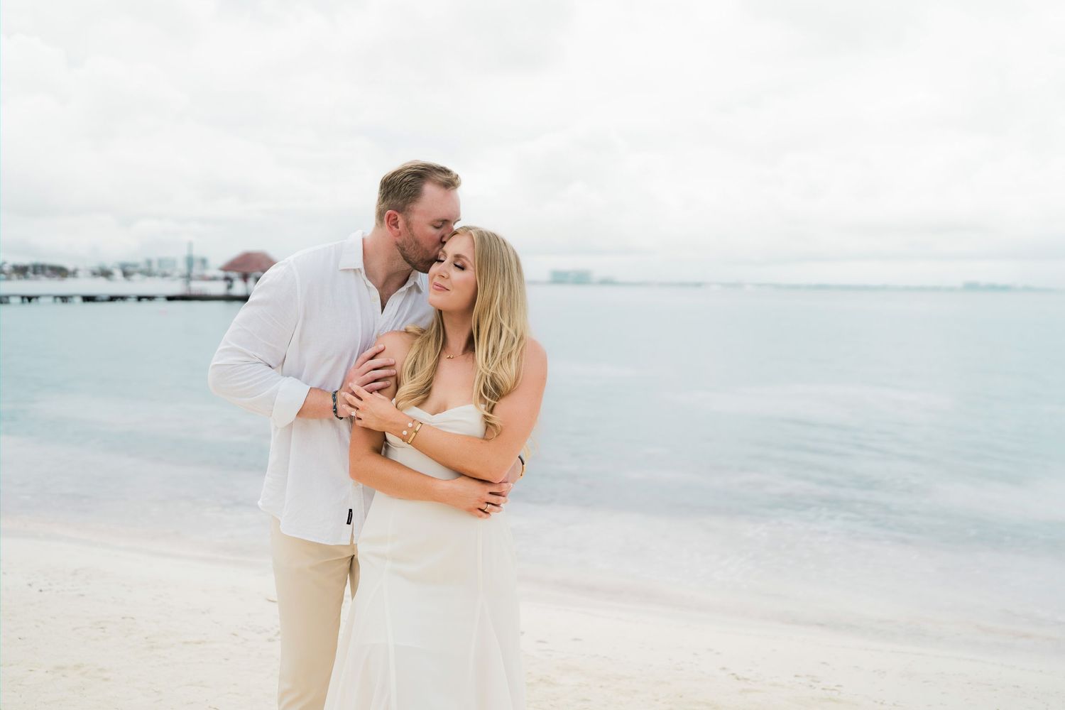 Cancun Engagement Photoshoot - Couple embracing on a tropical beach, man kissing woman's head, both in white attire with ocean backdrop.