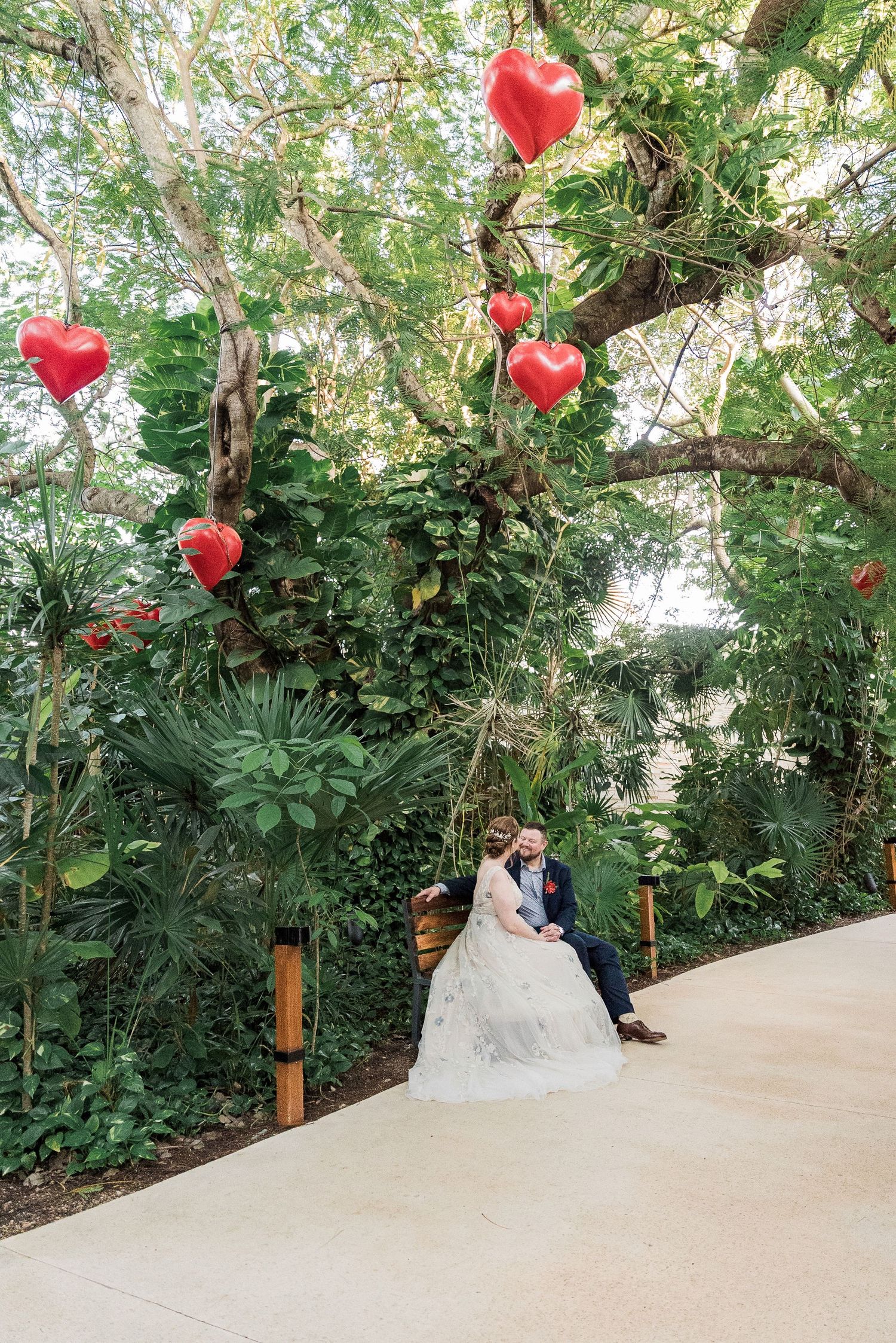 Secrets Akumal Wedding - Bride and groom sit on a swing under a lush tropical tree adorned with red heart balloons at an outdoor wedding.