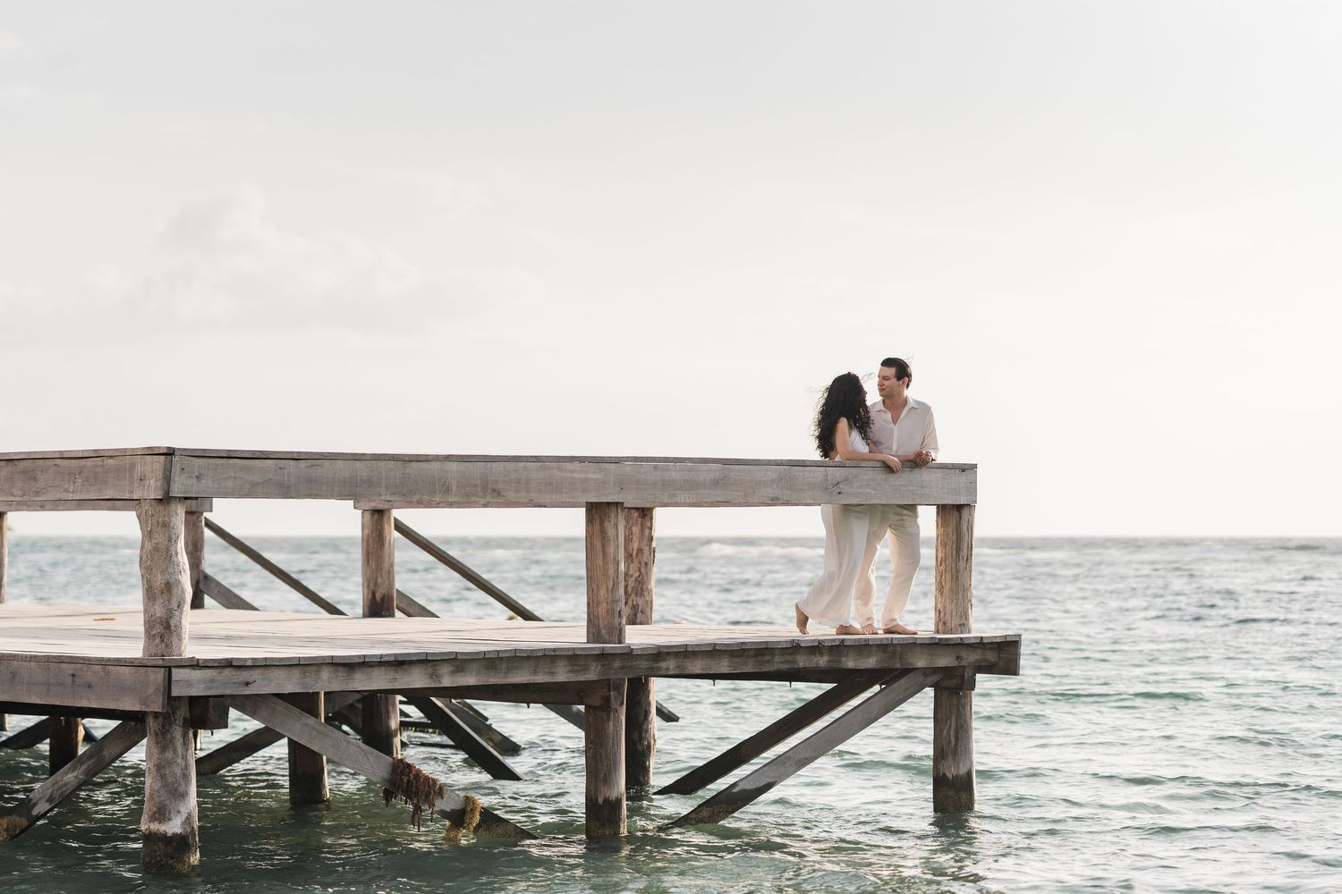 Couple embracing on a wooden pier over turquoise ocean waters, romantic seaside engagement photo.