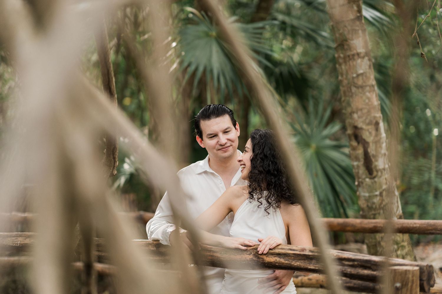 A couple shares a romantic moment on a wooden bridge surrounded by lush tropical trees and palm foliage.