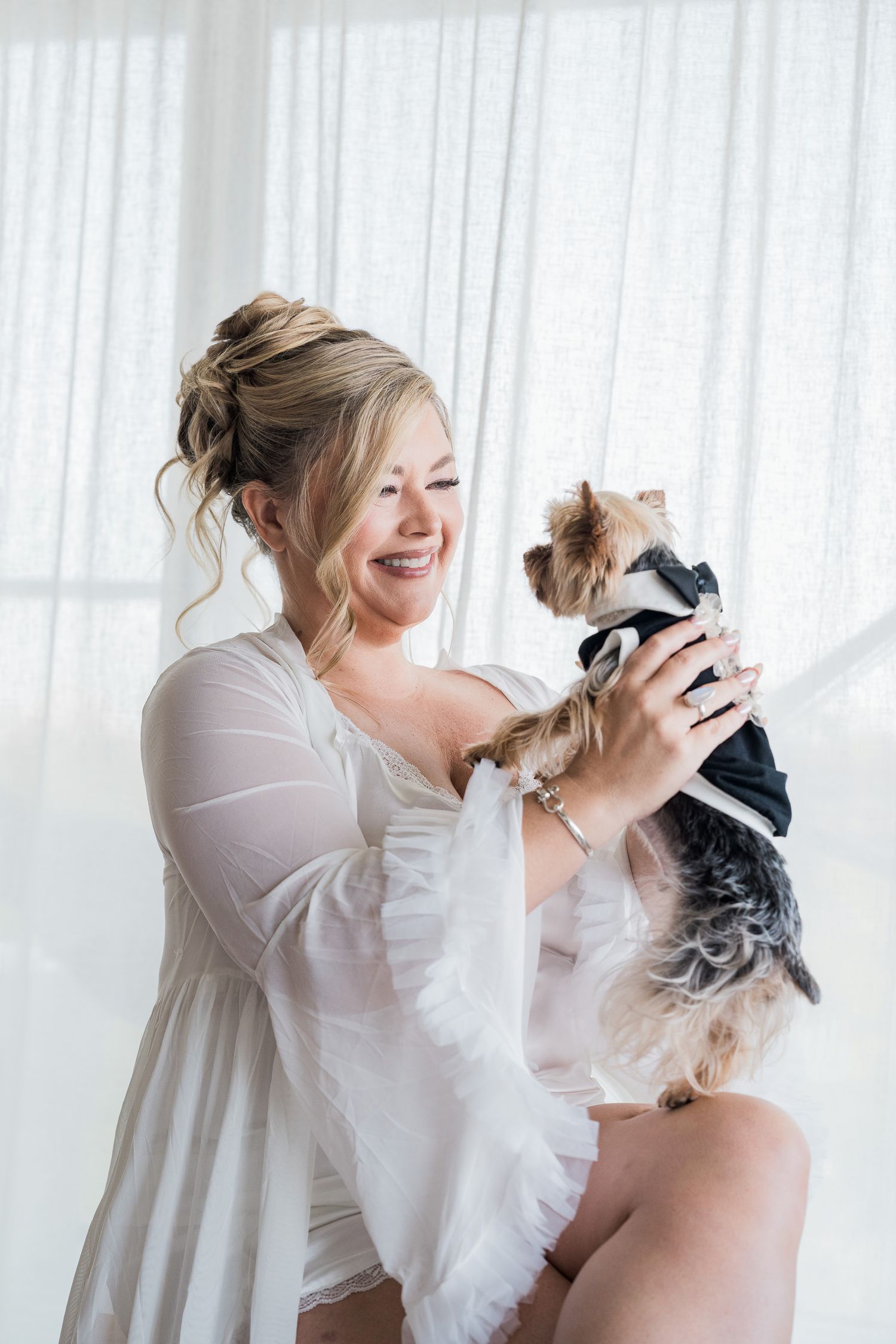 Hotel Xcaret México Wedding  - Smiling bride in white robe holds a Yorkshire Terrier wearing a black tuxedo near a bright window.
