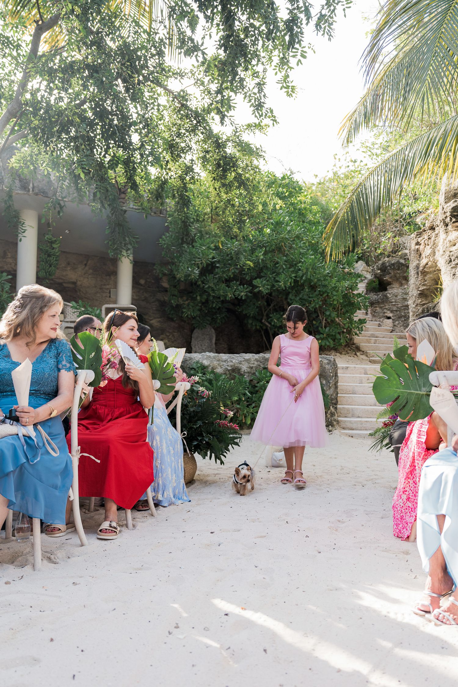 Outdoor wedding ceremony on sandy ground with guests in colorful dresses seated among tropical trees and ruins.