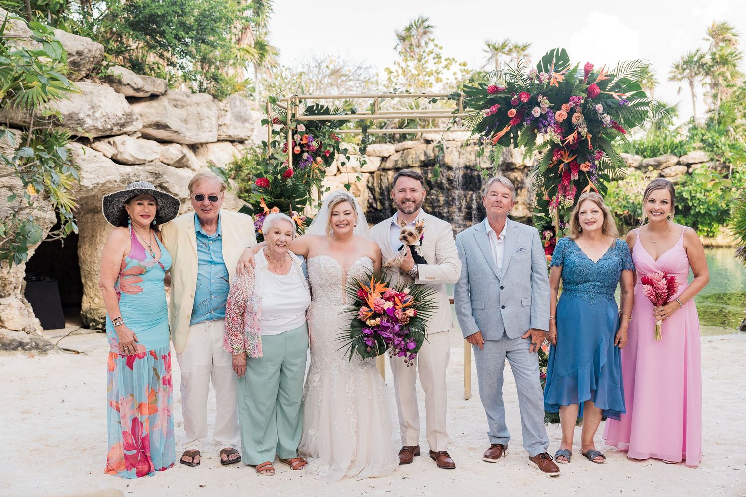 Hotel Xcaret México Wedding  - Wedding party poses before floral arch at tropical beach ceremony with bride holding colorful bouquet.