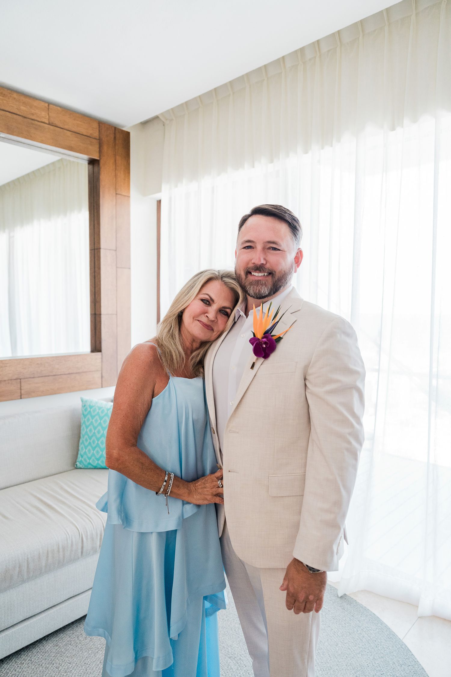 Smiling couple in wedding attire, man in beige suit with boutonniere, woman in light blue dress, posing indoors.