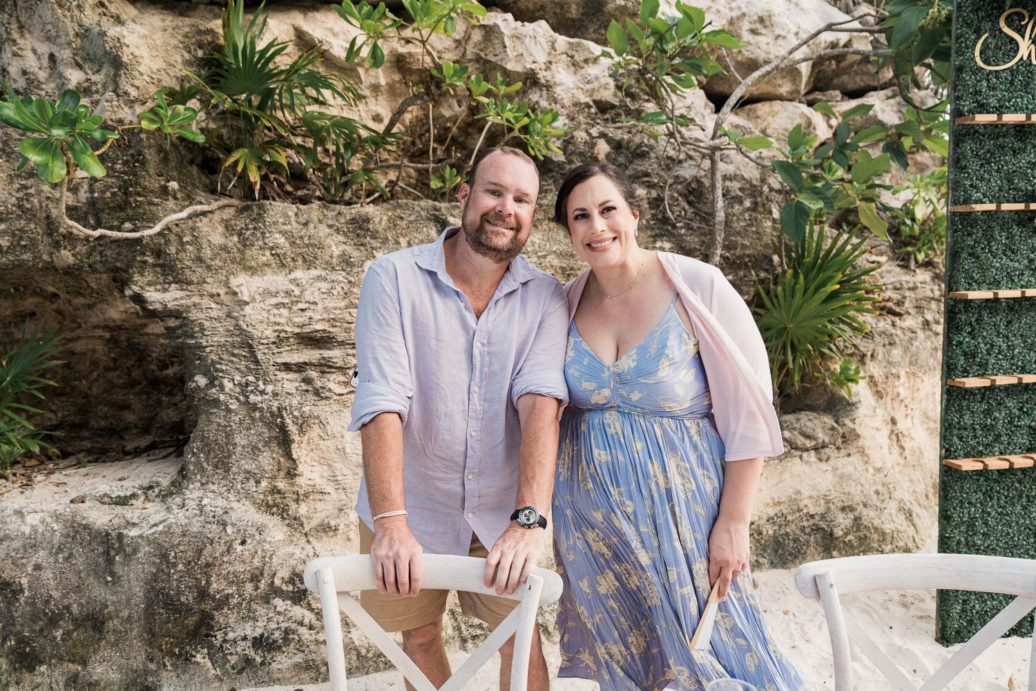 A smiling couple poses together outdoors near a stone wall with tropical greenery at a scenic vacation destination.