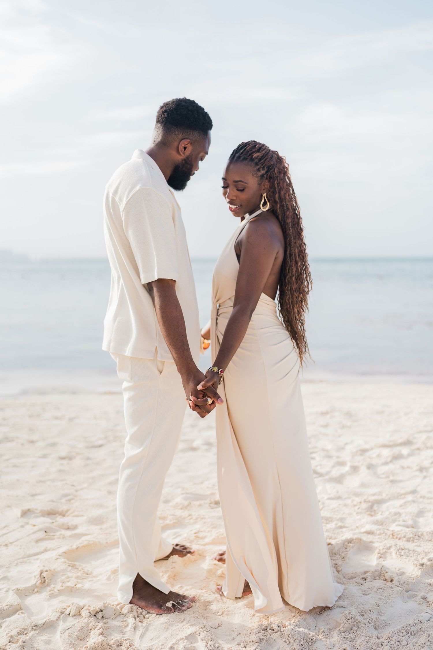A Black couple holds hands while standing barefoot on a white sandy beach, both dressed in matching cream-colored outfits.