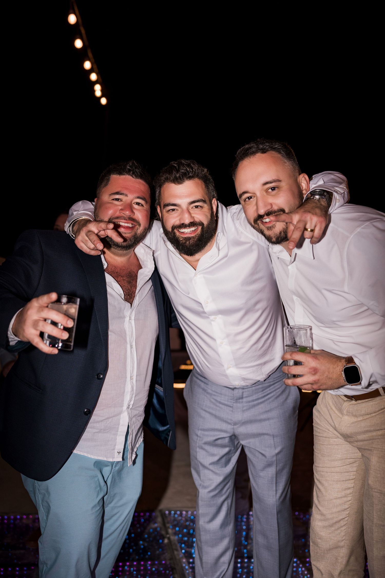 Three men smiling and posing together at a night event, holding drinks and cigars, dressed in smart casual attire.