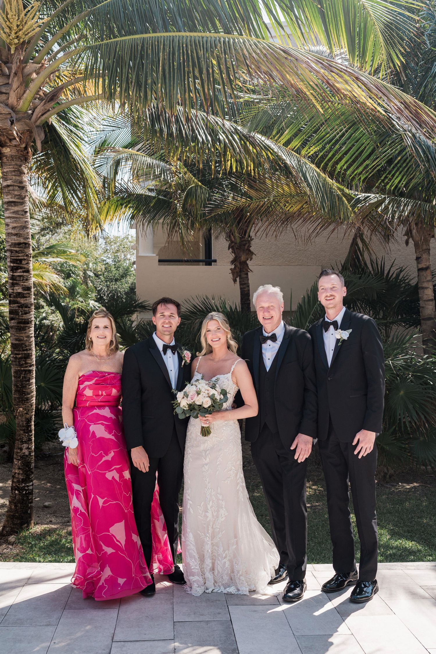 Wedding party posing under palm trees, bride in white gown with bouquet, groom and groomsmen in tuxedos, woman in pink floral gown.