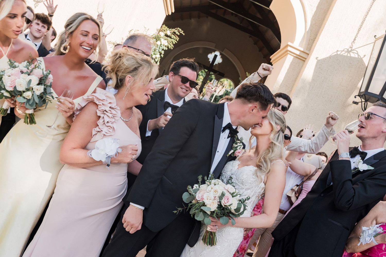 Bride and groom share a kiss surrounded by bridesmaids and guests outside a church after their wedding ceremony.