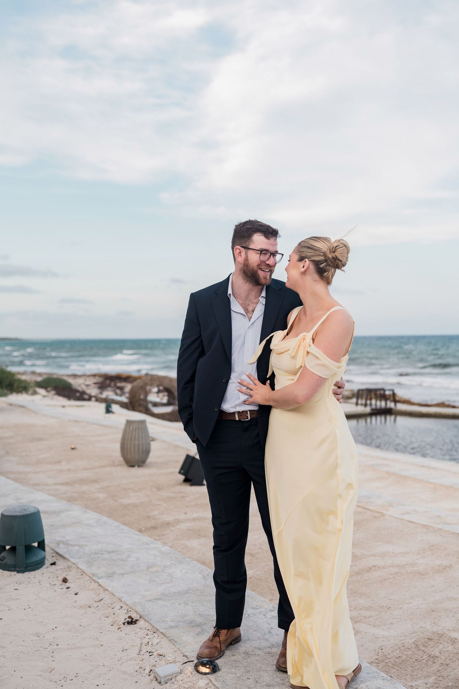 Couple in formal attire smiling at each other on a seaside terrace, man in suit, woman in yellow gown.