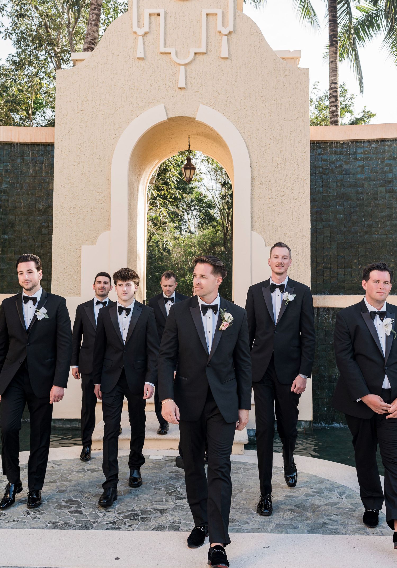 Groom and groomsmen in black tuxedos with bow ties walk confidently in front of an elegant arched venue.