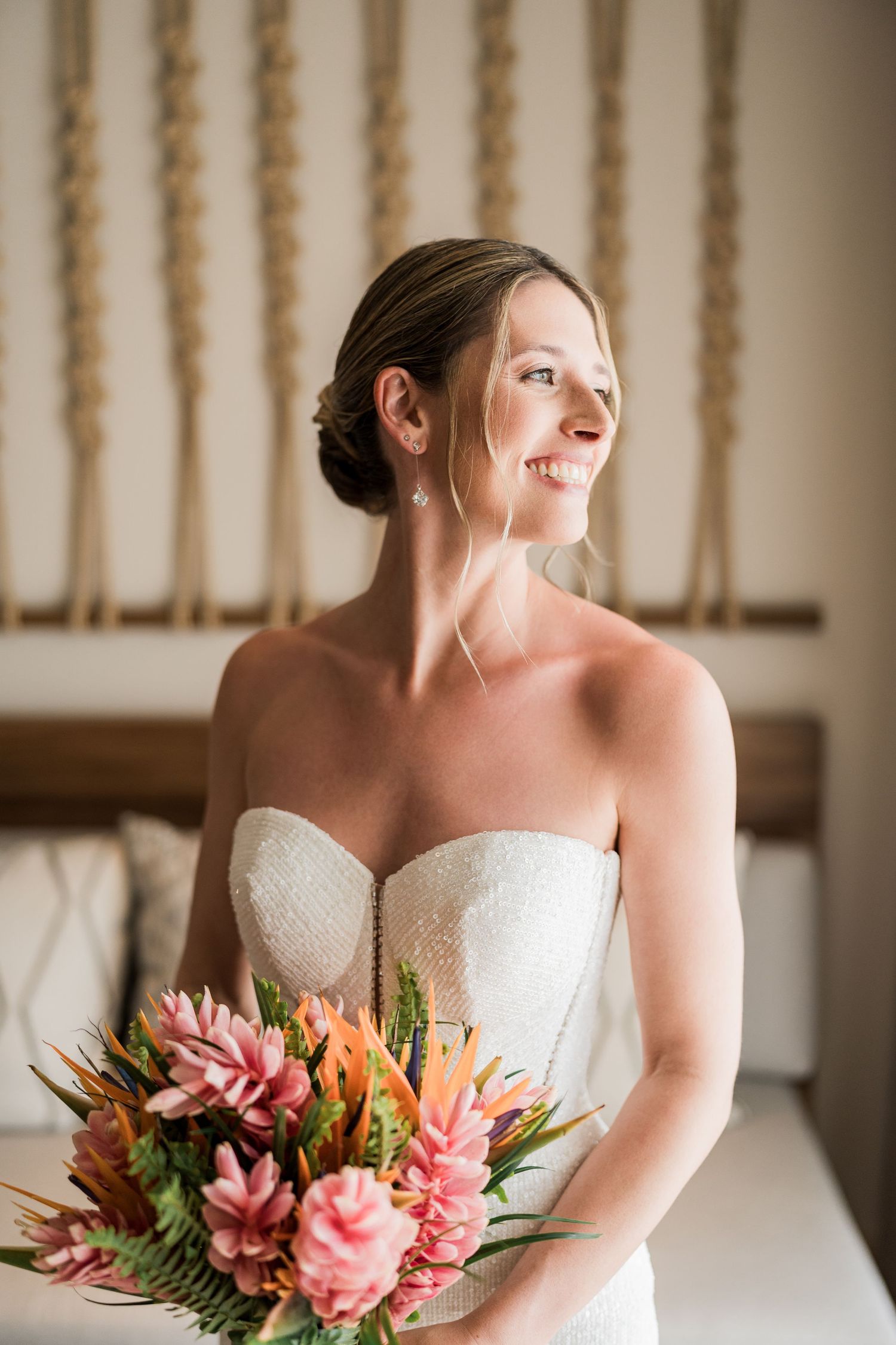 Smiling bride in strapless white gown holding tropical pink bouquet with ferns, posing indoors on her wedding day.