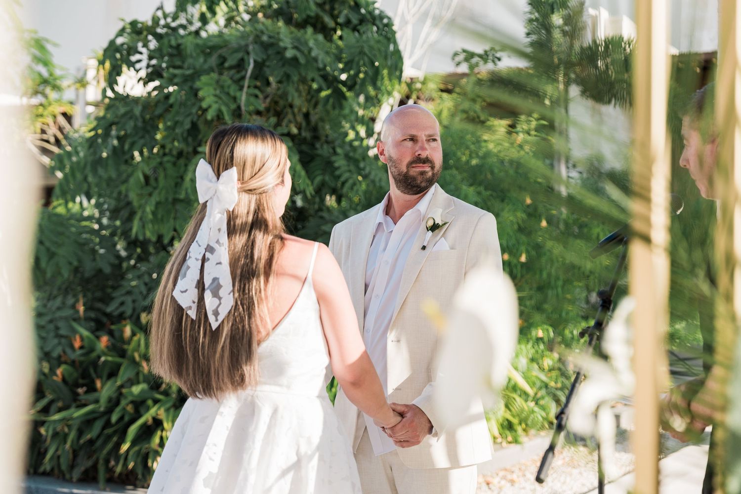 Groom in beige suit holds hands with bride in white dress during outdoor wedding ceremony.
