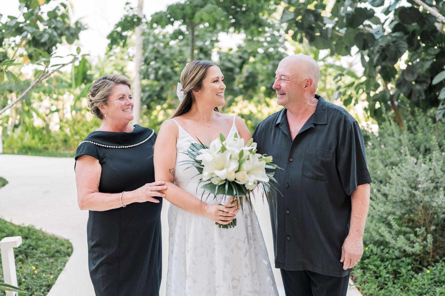 Bride in white gown holding bouquet smiles with two guests in black attire outdoors at a garden wedding venue.