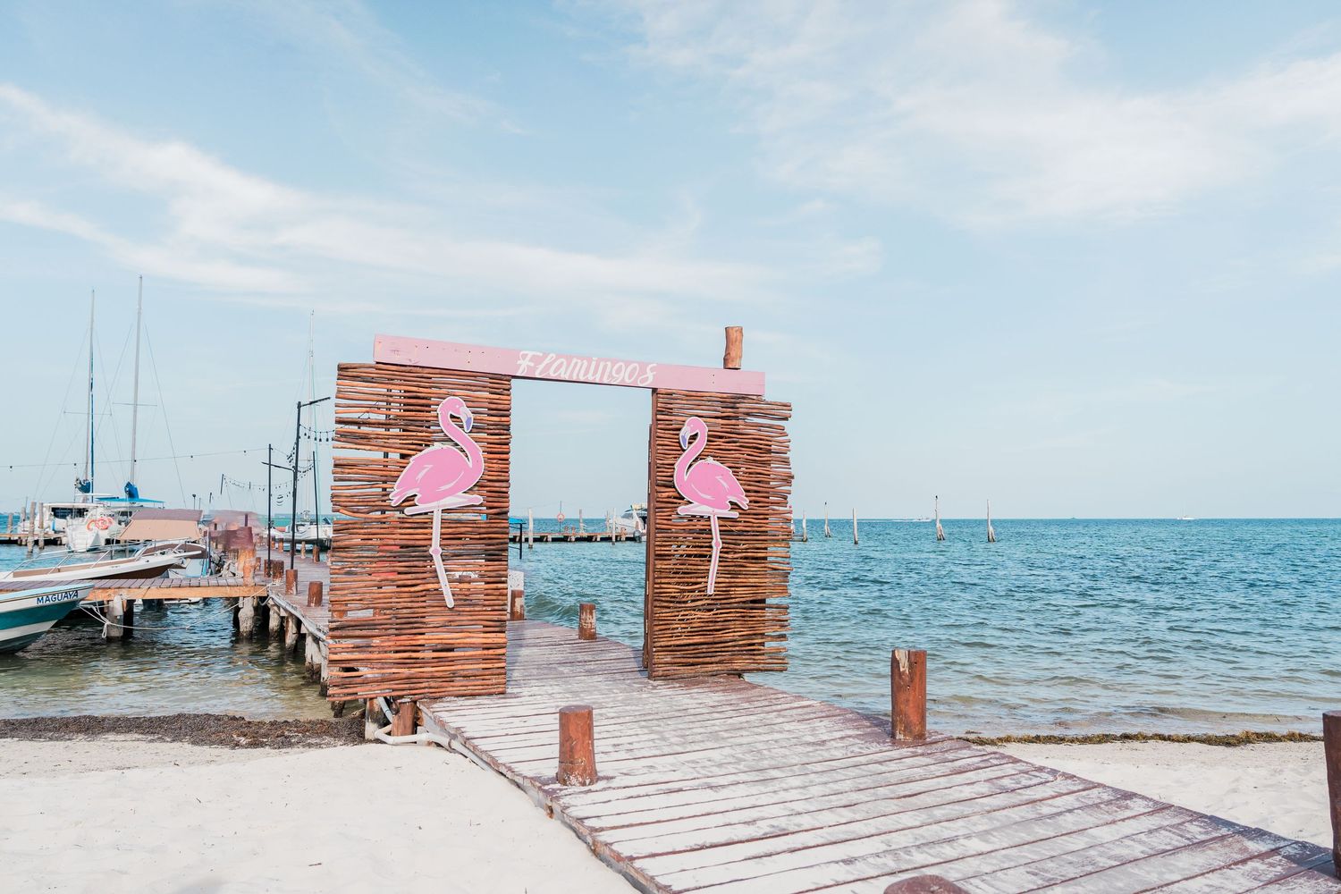 Wooden pier with pink flamingo decorations on a dock gate overlooking a calm turquoise sea under a blue sky.