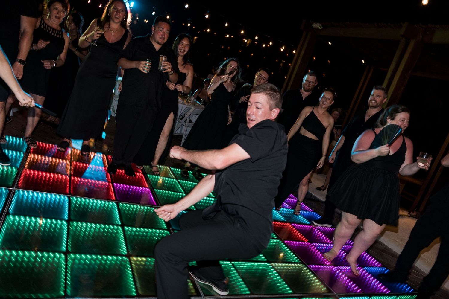 Man dancing energetically on a colorful LED light-up dance floor surrounded by guests at a party.