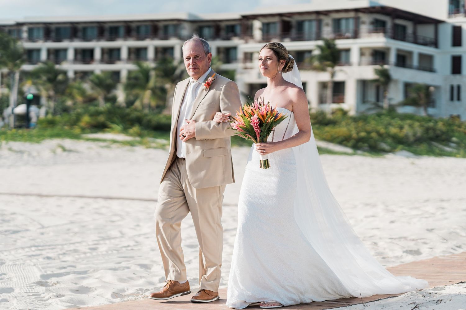 Bride in white gown holding colorful bouquet walks with older man in tan suit on sandy beach near resort.