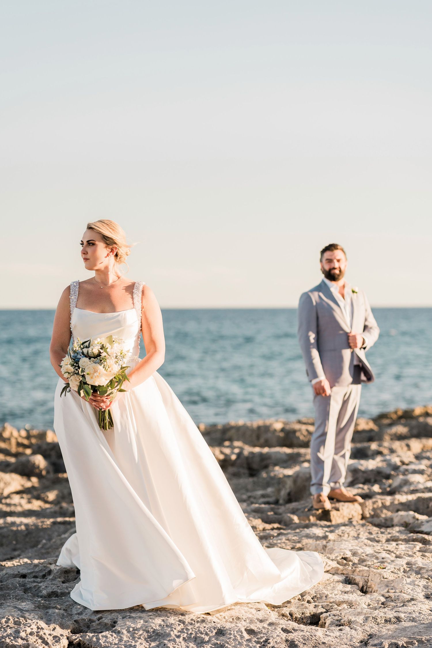 Bride in white gown holds bouquet on rocky shore, groom in gray suit stands behind with ocean backdrop. Hard Rock Riviera Maya Wedding