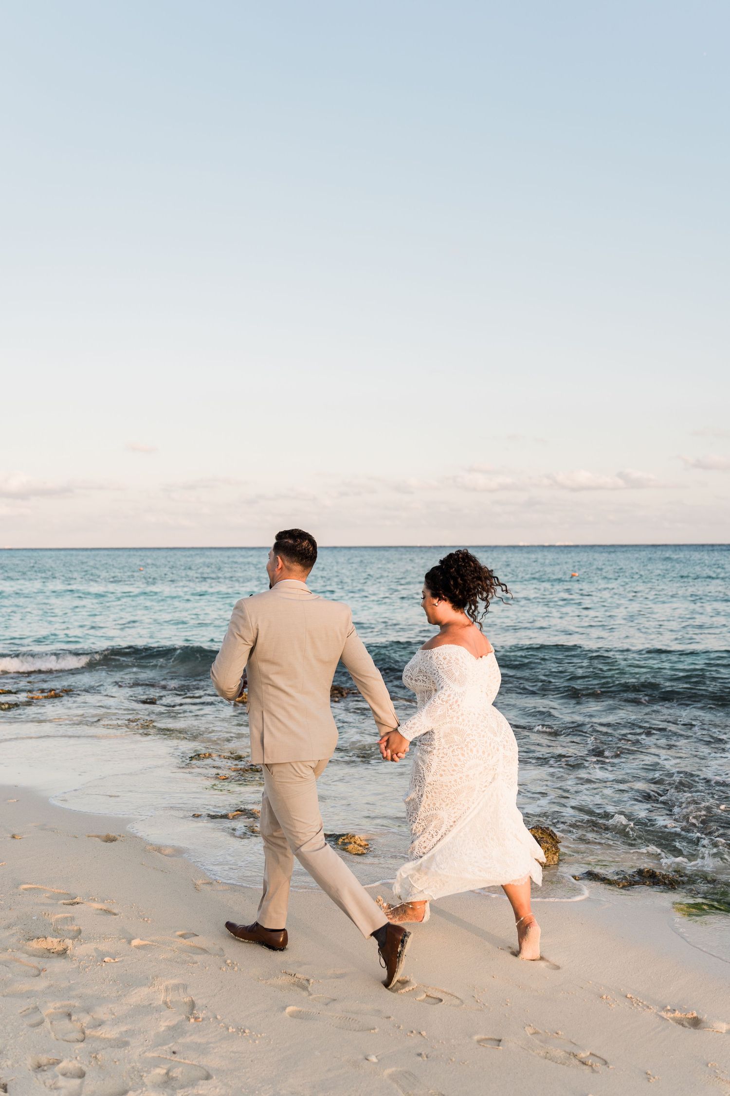 Xcaret Arte Wedding - Couple in wedding attire holding hands and walking along a sandy beach at sunset with turquoise ocean waves.