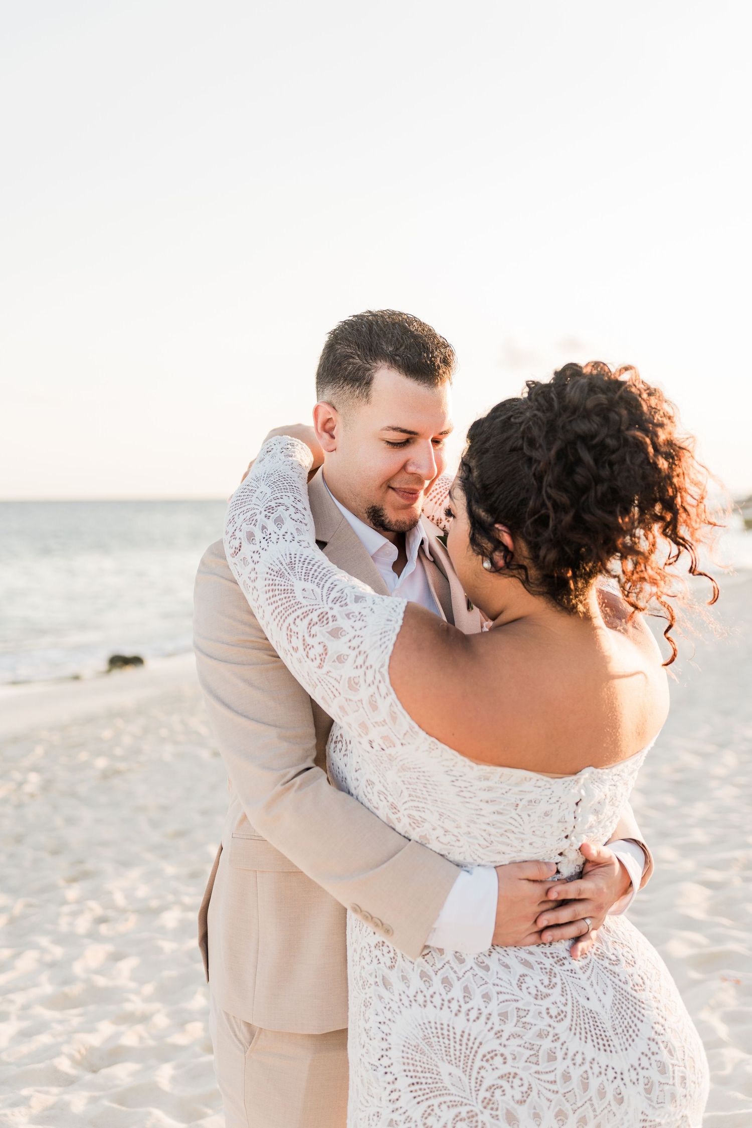 Xcaret Arte Wedding - Bride and groom embracing on a sandy beach at sunset, bride in lace off-shoulder gown, groom in tan suit.