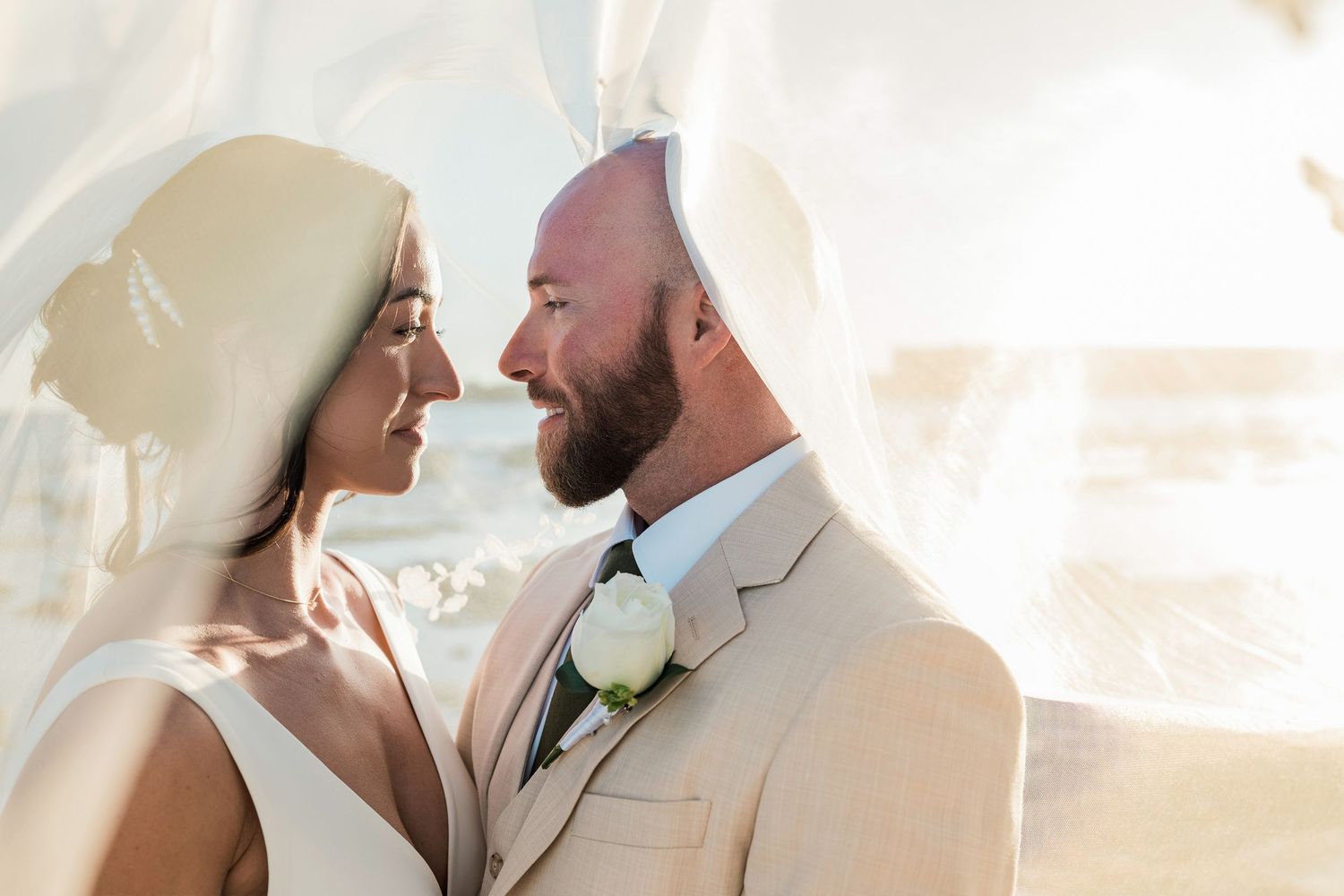 Bride and groom share an intimate moment under a flowing veil at an outdoor wedding ceremony.
