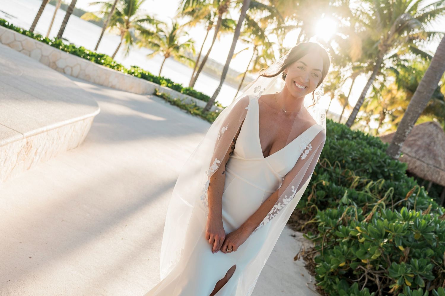 Bride in white gown with flowing veil smiles outdoors near palm trees and beach at golden hour.
