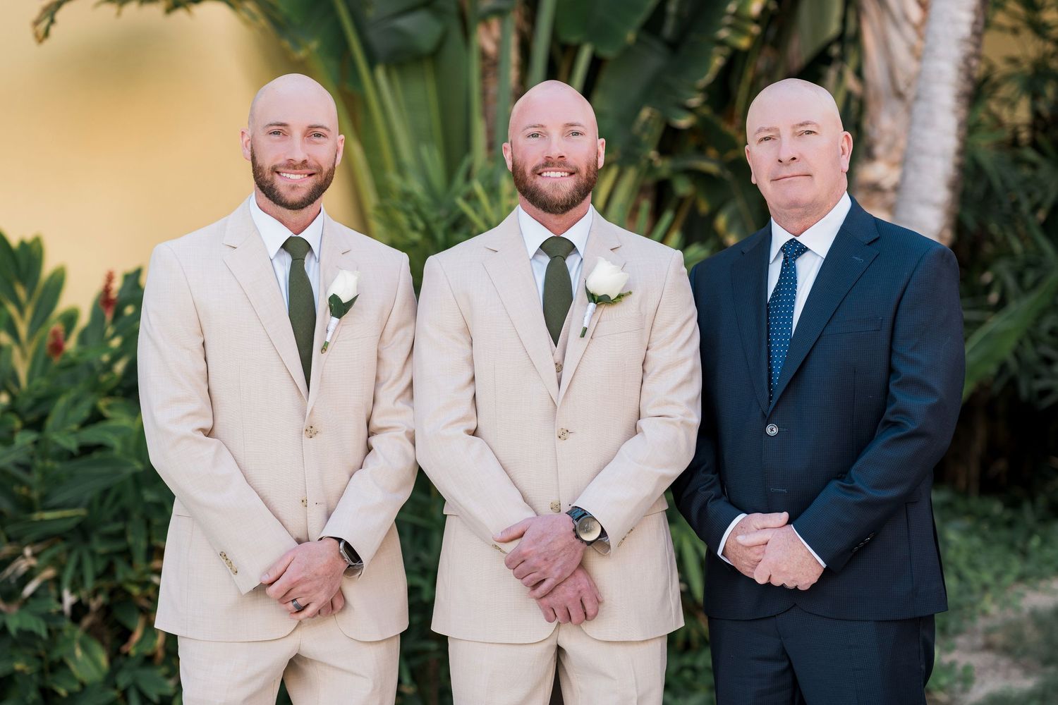 Three men in formal attire, two in tan suits with boutonnieres and one in navy, posing outdoors at a wedding.