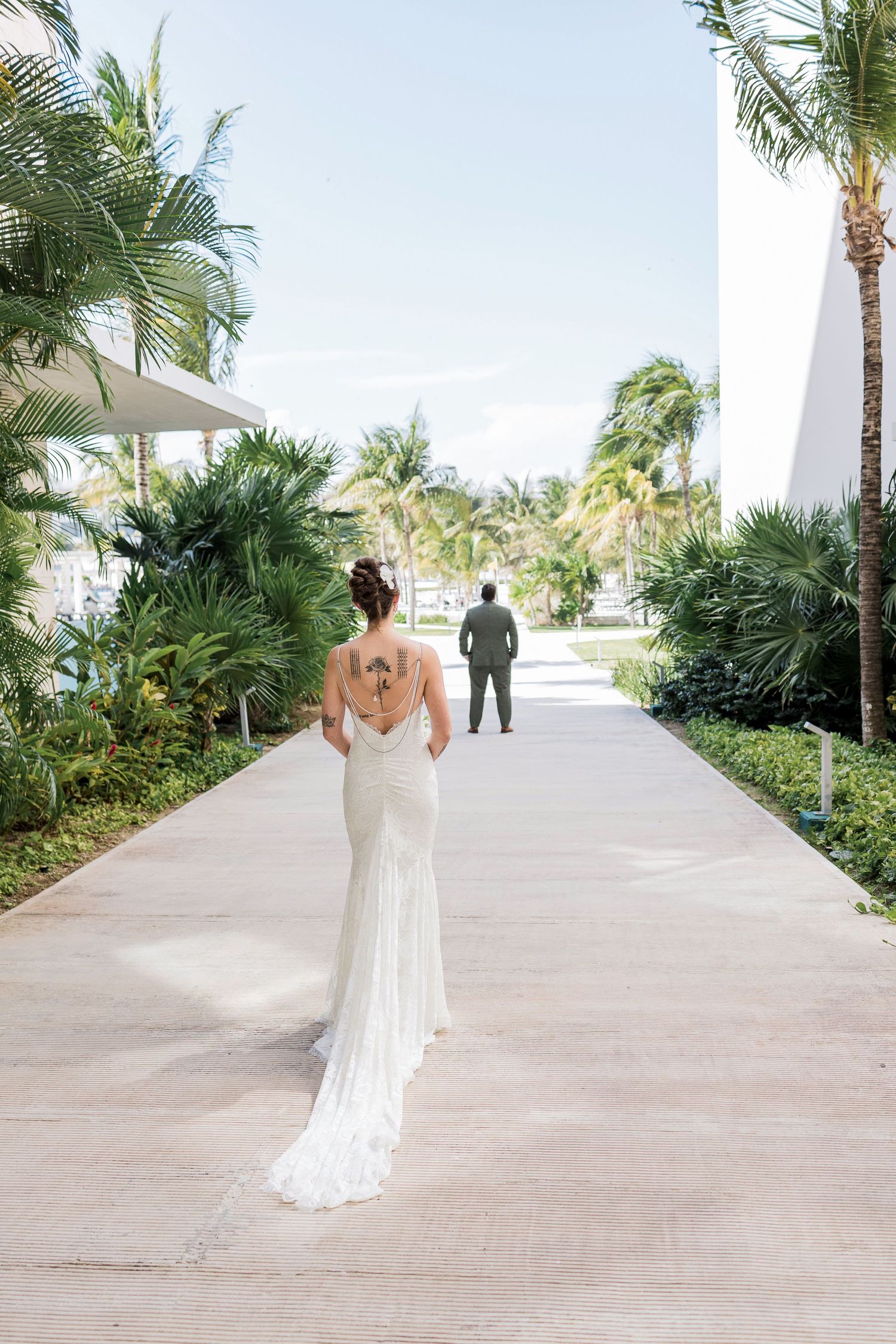 Bride in backless wedding gown with tattoo walks toward groom on tropical resort pathway lined with palm trees.