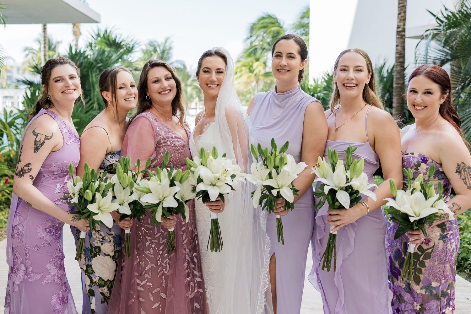 Bride in white gown with bridesmaids in purple dresses holding white lily bouquets outdoors at a tropical wedding.