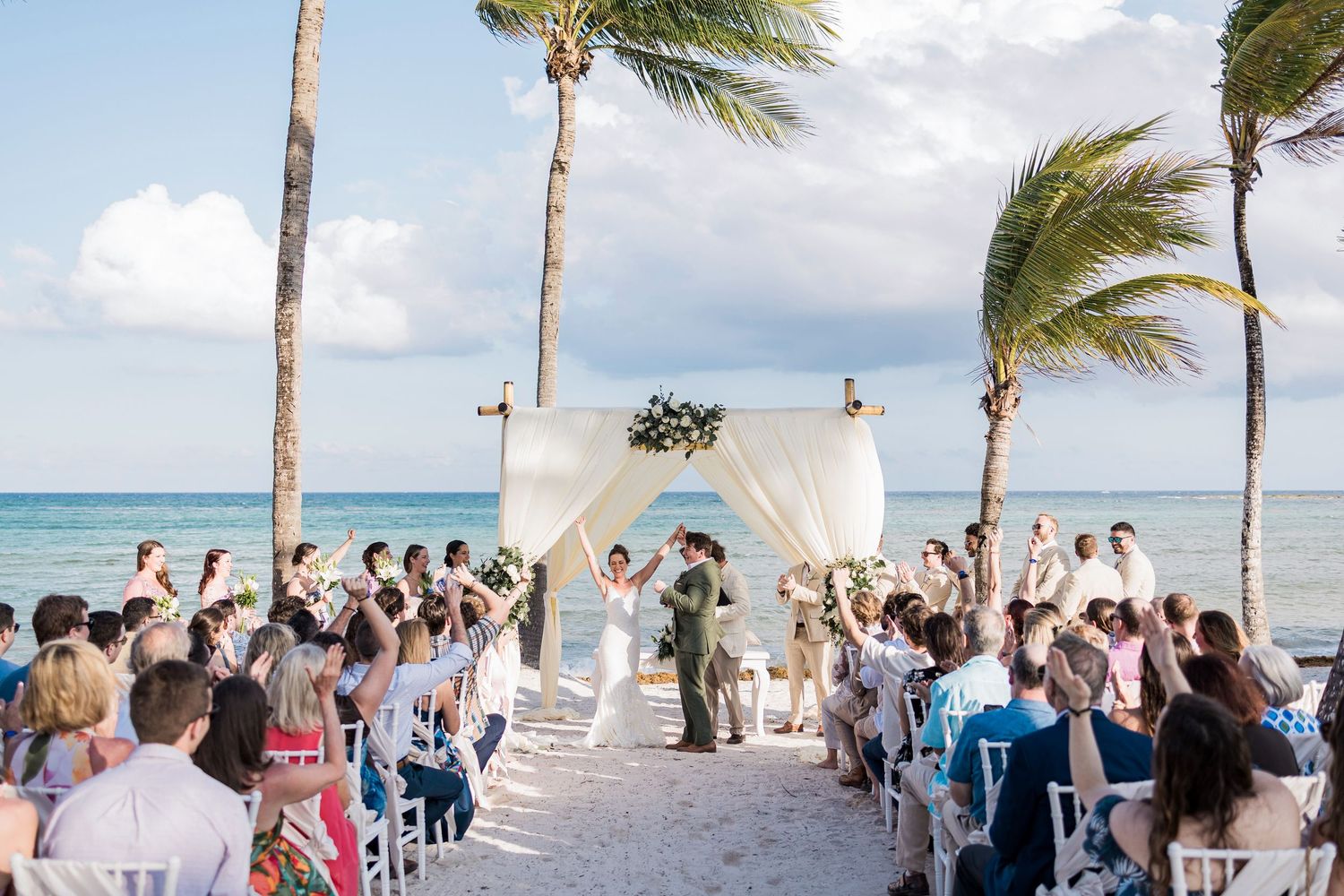 Romantic beach wedding ceremony with white drape altar, palm trees, and ocean backdrop with seated guests.