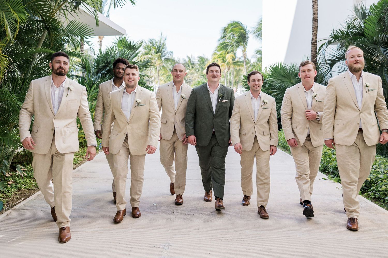 Groom in olive suit walks with seven groomsmen in tan suits along outdoor pathway at tropical wedding venue.