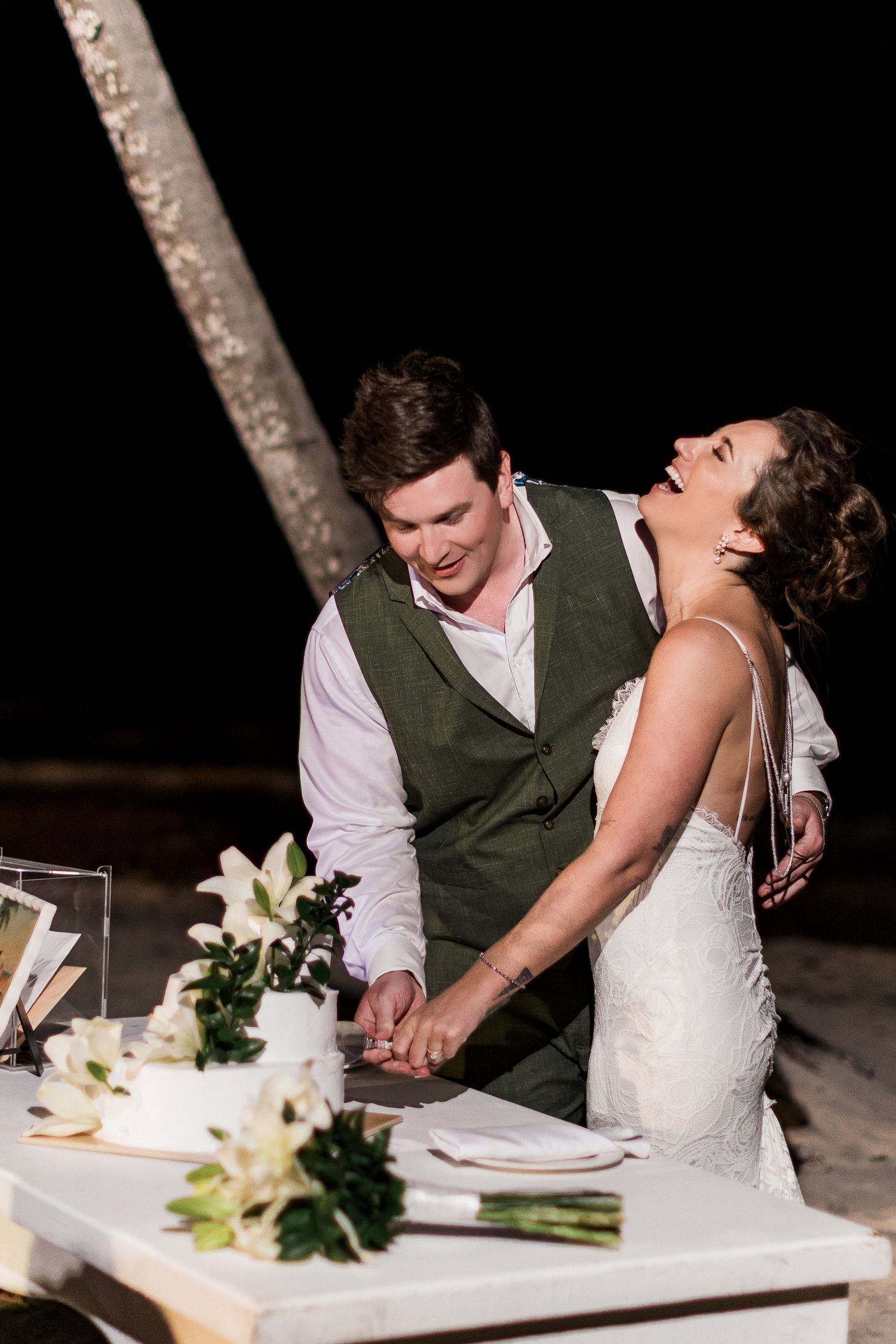 Bride and groom laughing while cutting white wedding cake with lilies at outdoor beach night reception.
