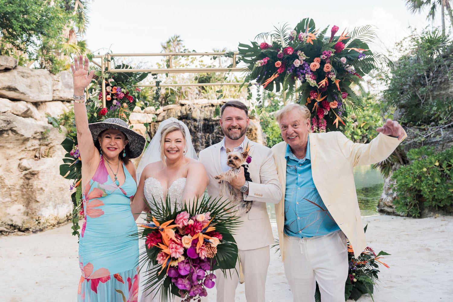 Tropical beach wedding ceremony with bride and groom celebrating with guests under colorful floral arch.