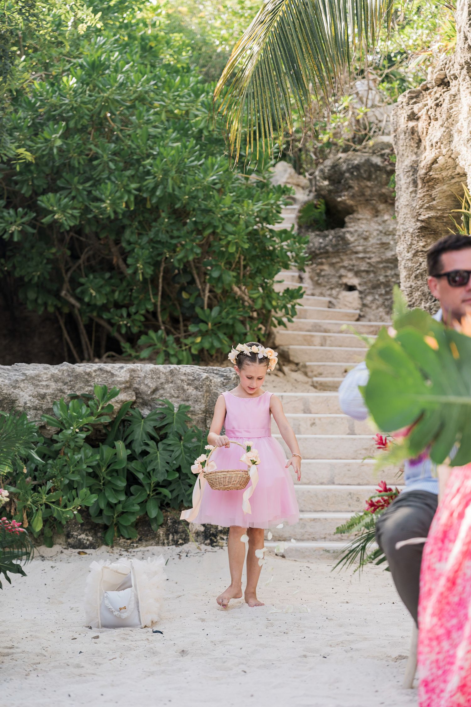 Hotel Xcaret México Wedding  - Young flower girl in pink dress and floral crown walks barefoot on sandy path at outdoor beach wedding.