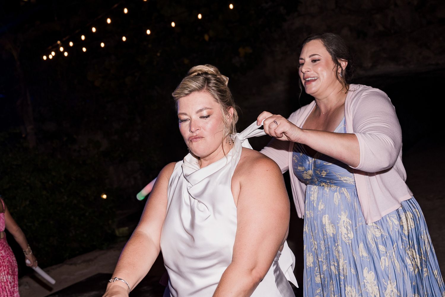 Bridesmaid in pink cardigan adjusting bride's white halter dress during outdoor evening wedding reception.