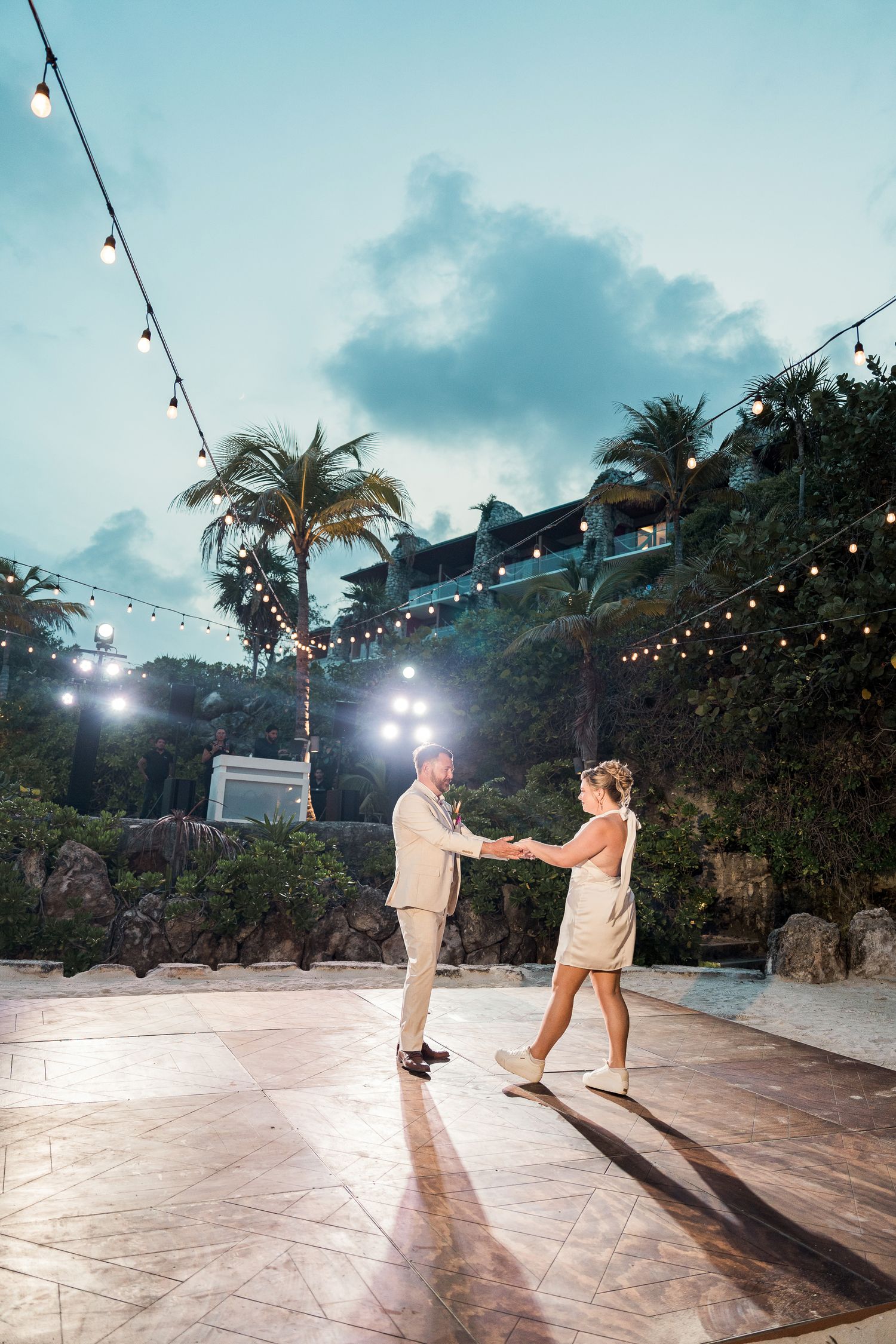 Couple sharing first dance on outdoor patio with string lights and tropical palm trees at dusk.