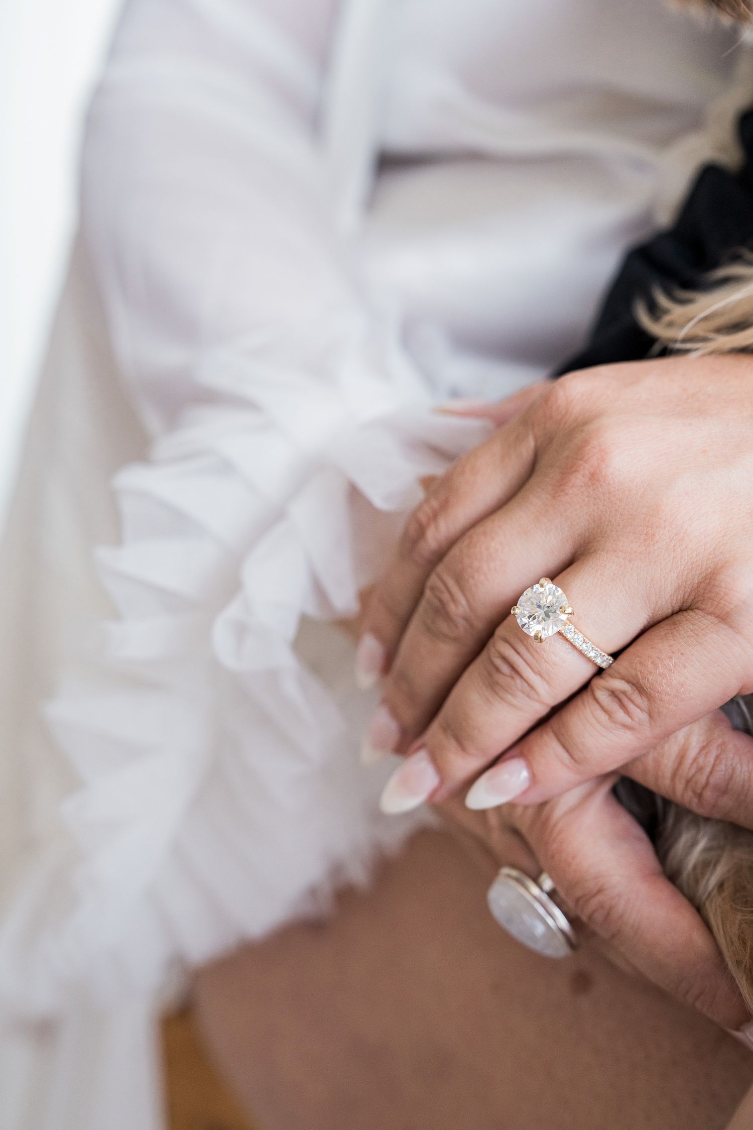Close-up of a bride's hand wearing a stunning diamond halo engagement ring, resting on a white ruffled wedding dress.