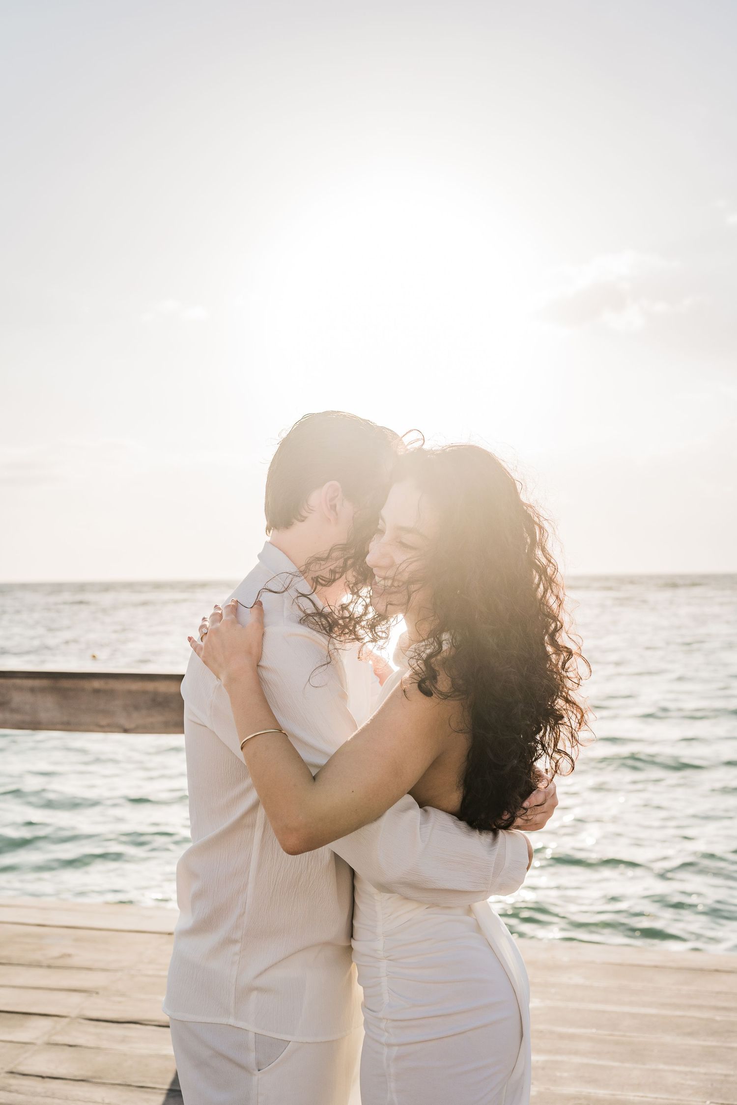 Couple embracing on a sunlit pier with ocean backdrop, wearing white outfits during a romantic beach engagement session.