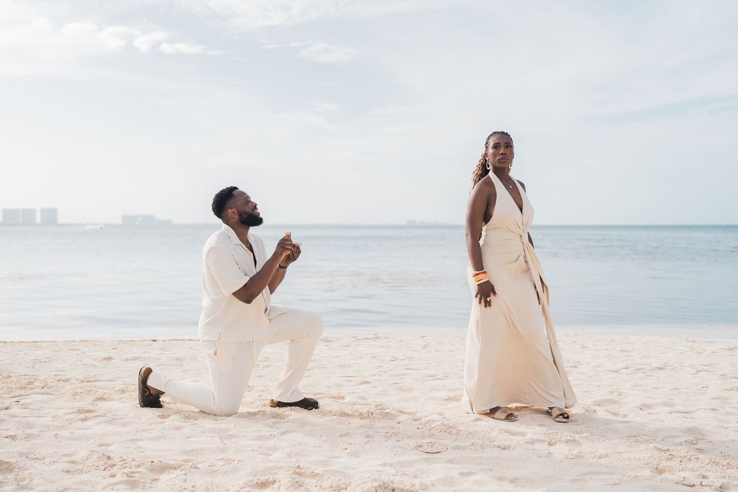 Man proposes on one knee to woman in white dress on sandy beach with ocean backdrop.
