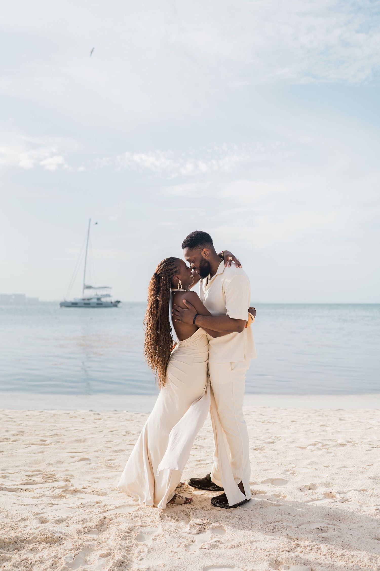 A Black couple shares a romantic kiss on a sandy beach, both dressed in elegant white outfits with a sailboat in the background.