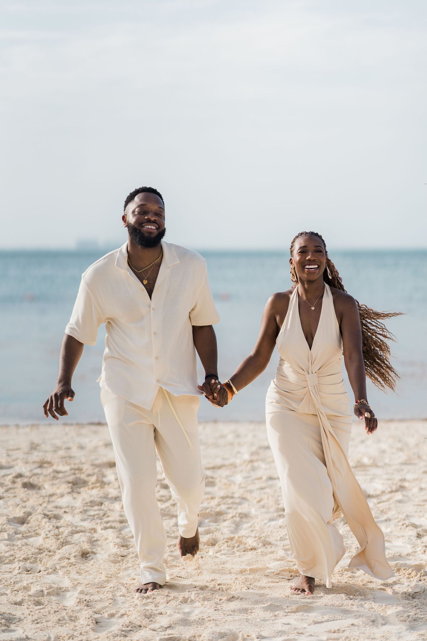 A joyful Black couple in matching cream outfits holds hands while walking barefoot on a sandy beach.